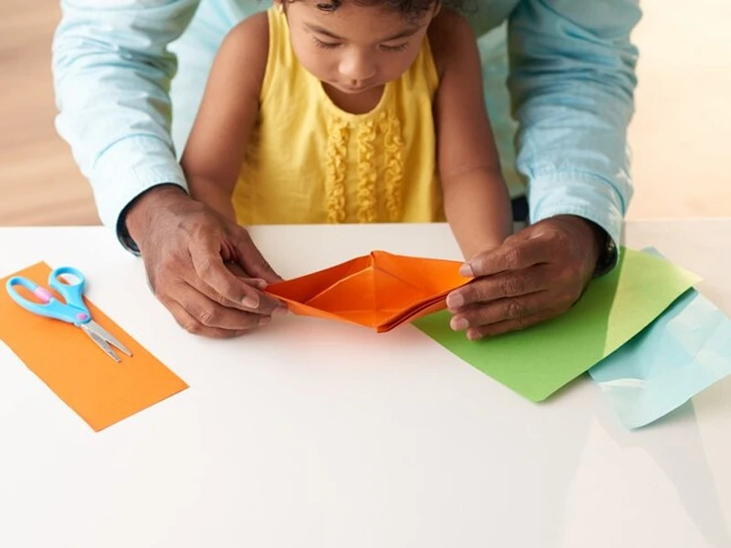 Parent helping child fold origami paper during a hands-on craft activity with colourful sheets and scissors on the table
